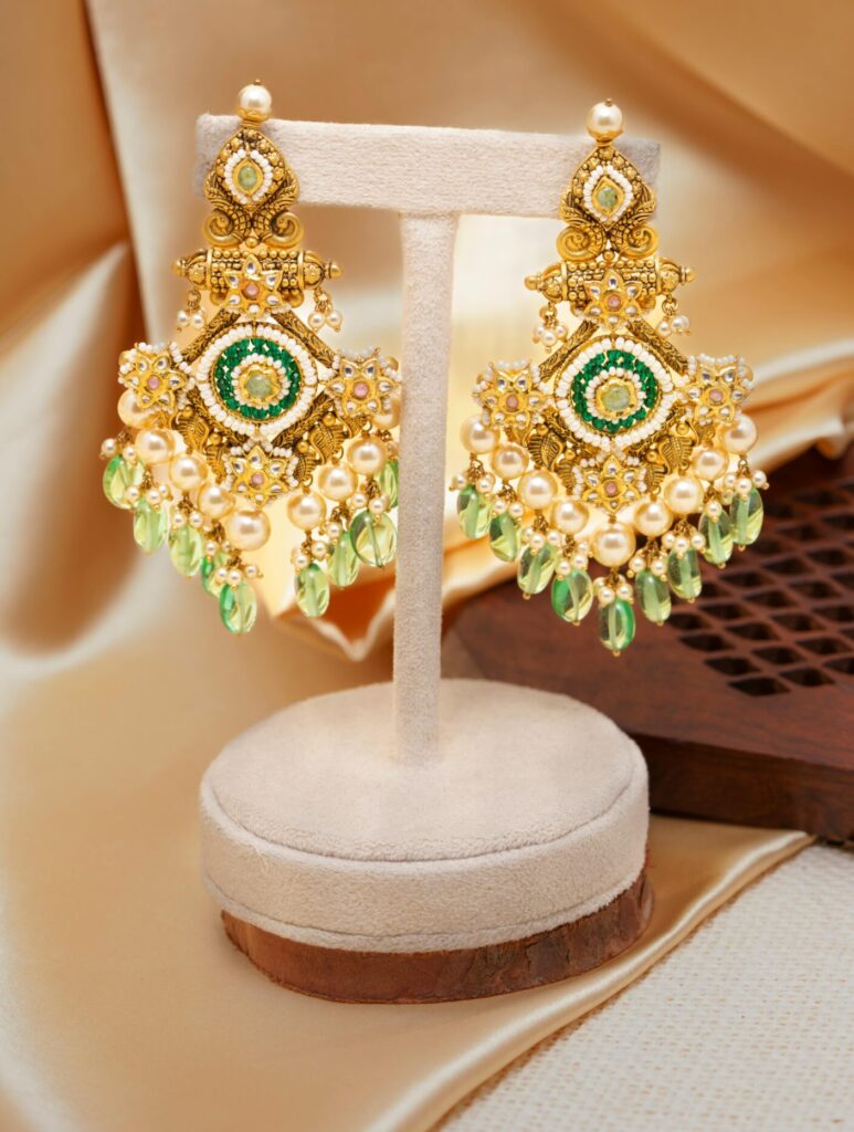 Close-up of luxurious gold chandelier earrings with green gems on a beige background.